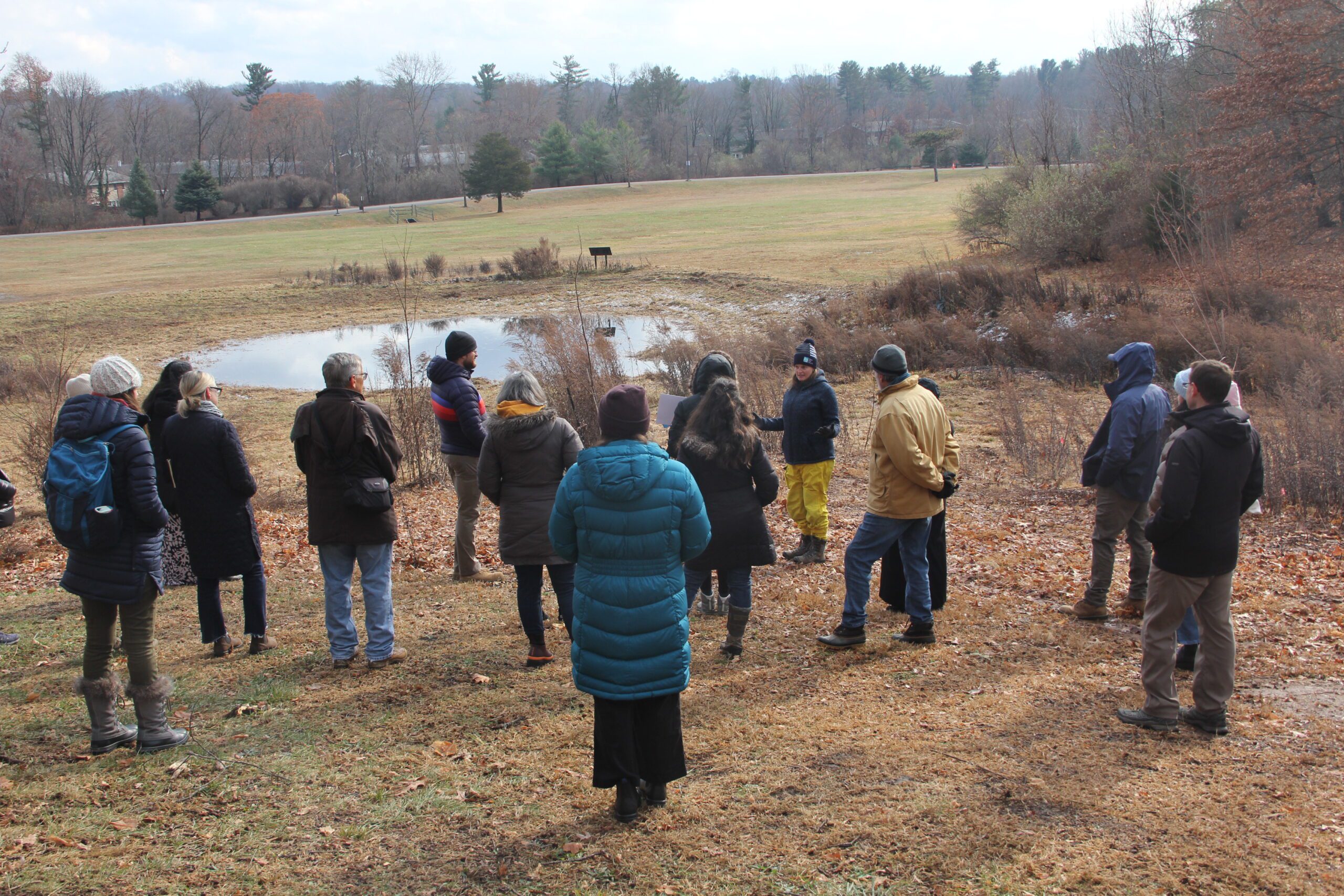 A group of people dressed in warm coats and hats stand on a grassy hillside overlooking a small pond and field. There are trees in the distance.
