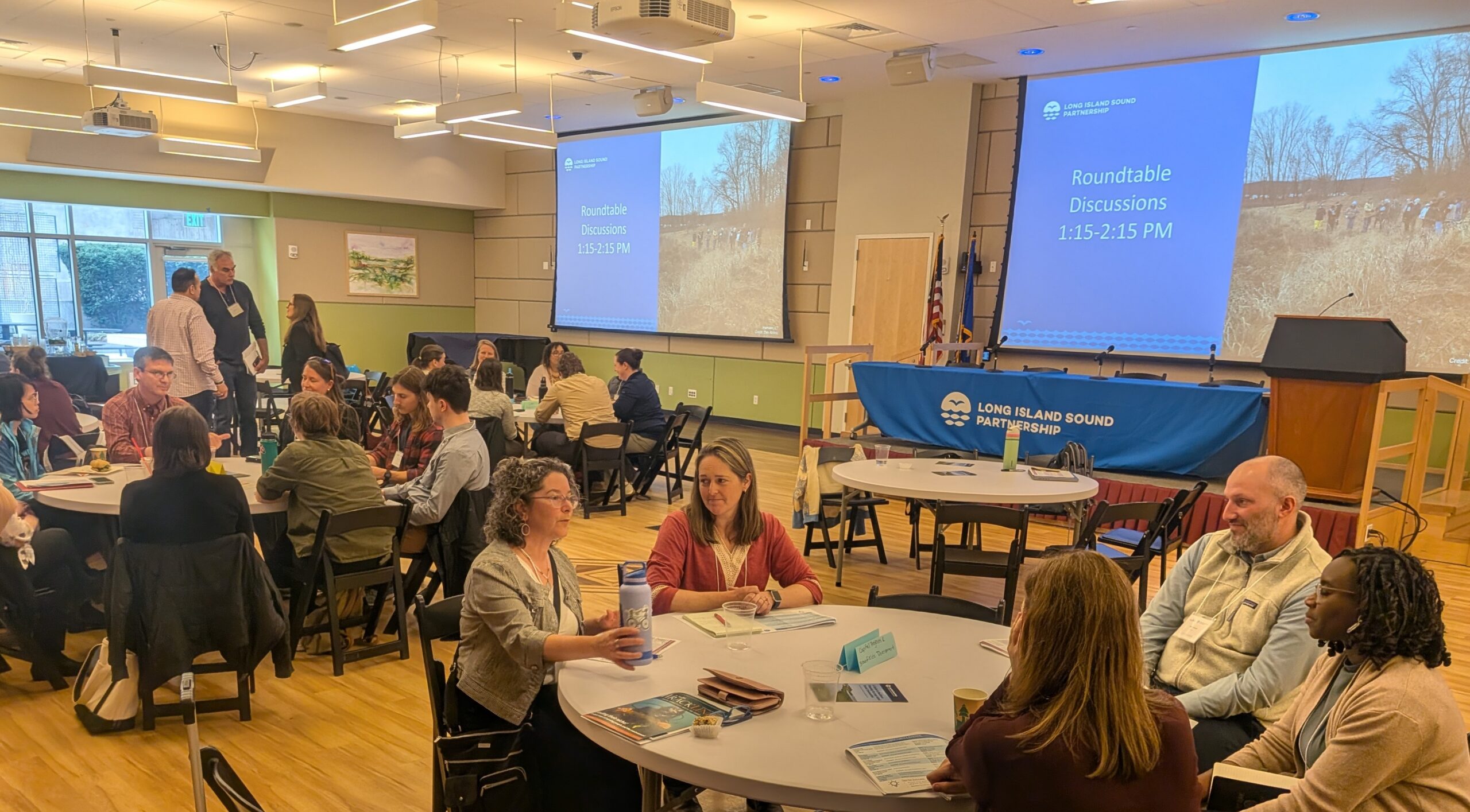 People seated around 3 round tables in a large room, talking with each other. At the  front of the room are two presentation screens, and a table, and podium on a small stage.