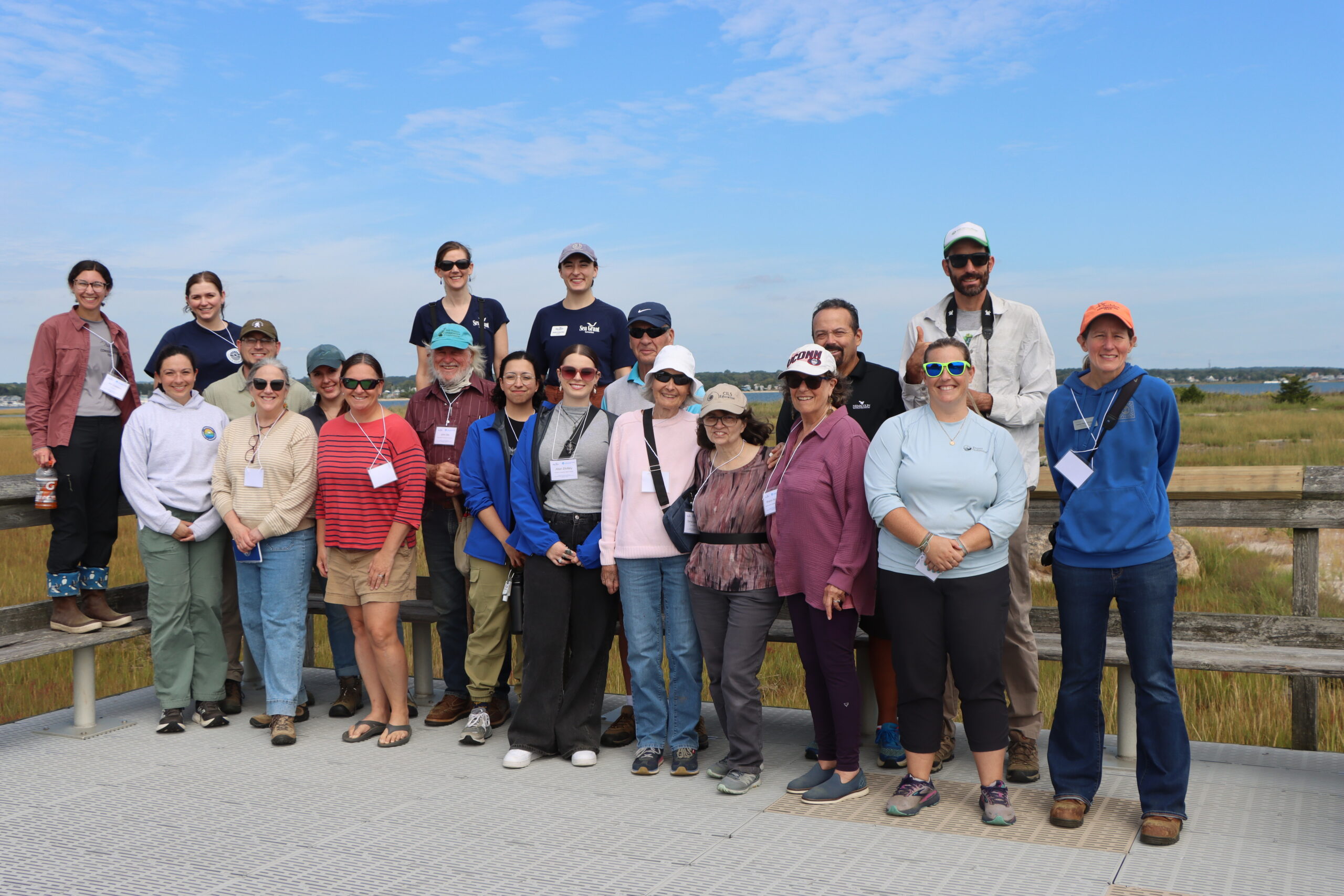 20 people standing outside on marsh viewing platform posed for picture under blue sky. Green marsh grasses are in sight behind group.