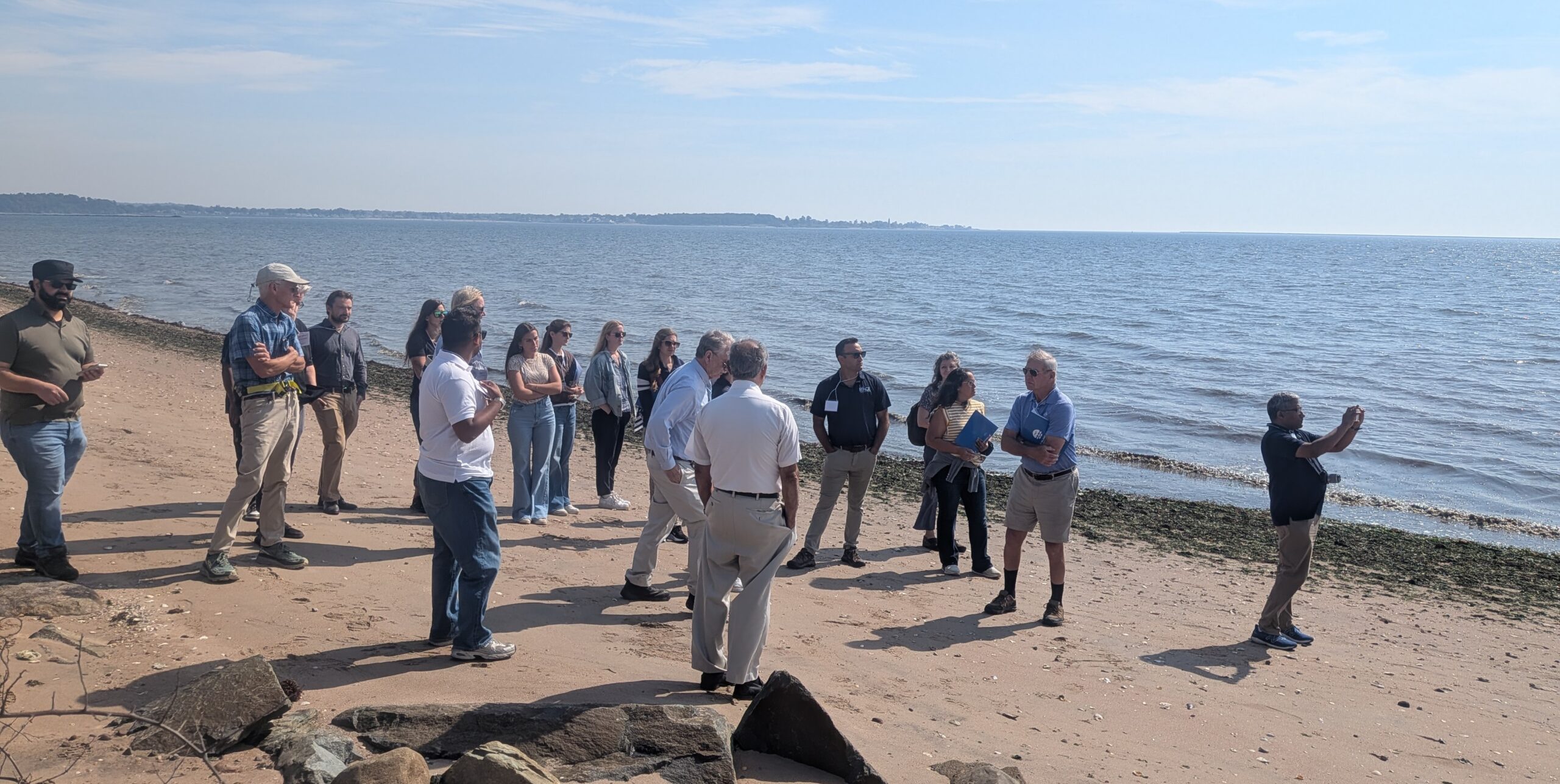 A group of people standing on a sandy beach close to the shoreline. Most are in casual work attire, and they appear to be listening to a speaker. The water is calm and there is land in the distance.